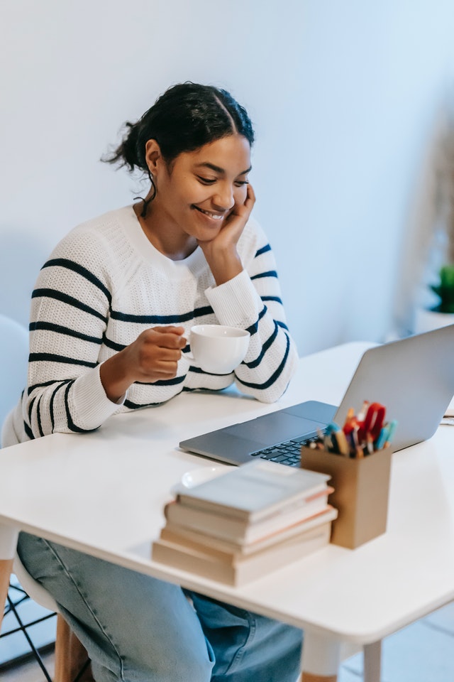 A woman in a white and navy striped sweater sits at a desk, smiling as she looks at her laptop while holding a cup of coffee. The desk also has a stack of books and a container filled with colorful pens and pencils.