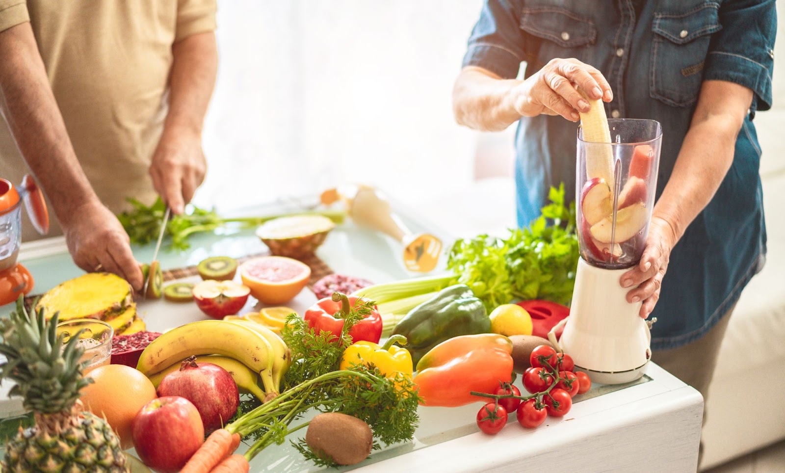 A senior couple in the kitchen prepare a smoothie in a blender with various fruits and vegetables laid out on a table.