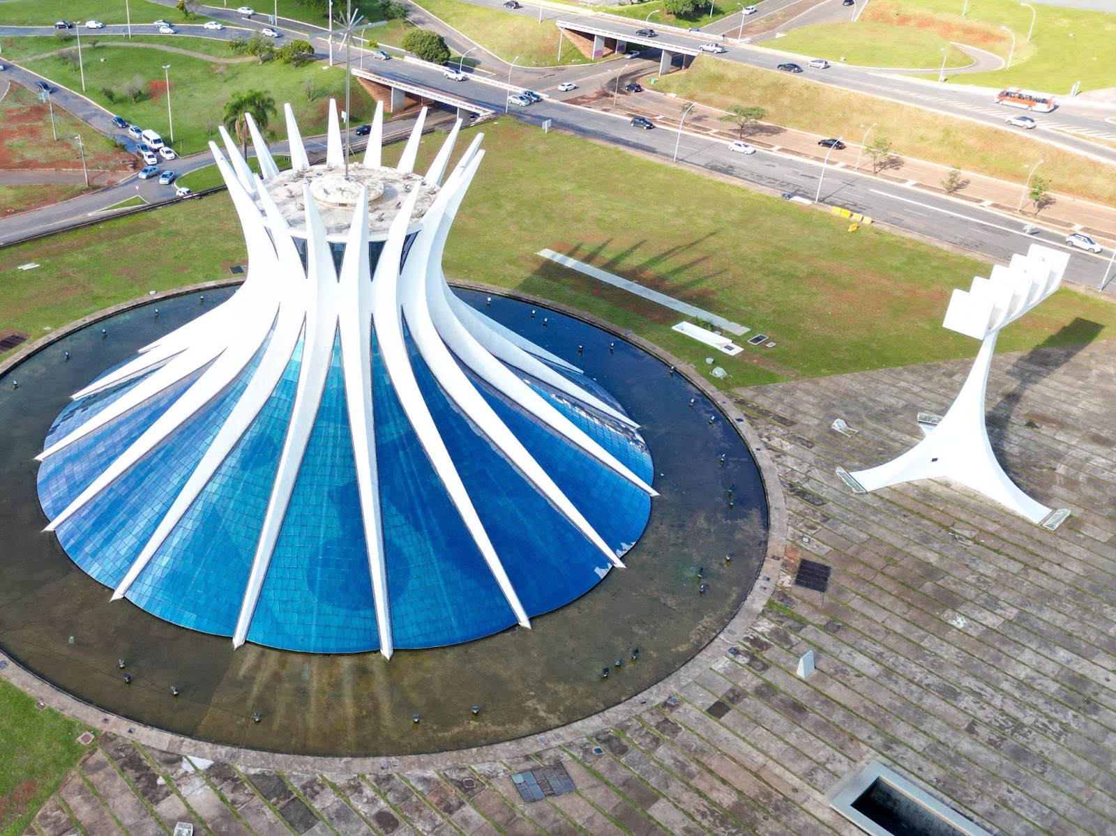 Catedral de Brasília vista de cima. O templo é rodeado por um espelho d’água em formato circular. À direita da entrada, aparece o campanário, estrutura similar a uma torre, onde ficam quatro sinos enfileirados.
