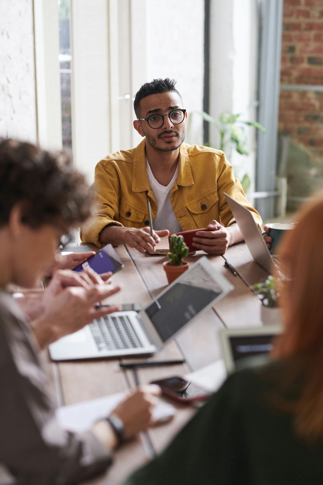 A man in a yellow shirt sits at a table during a meeting, holding a phone and listening, while others use laptops.