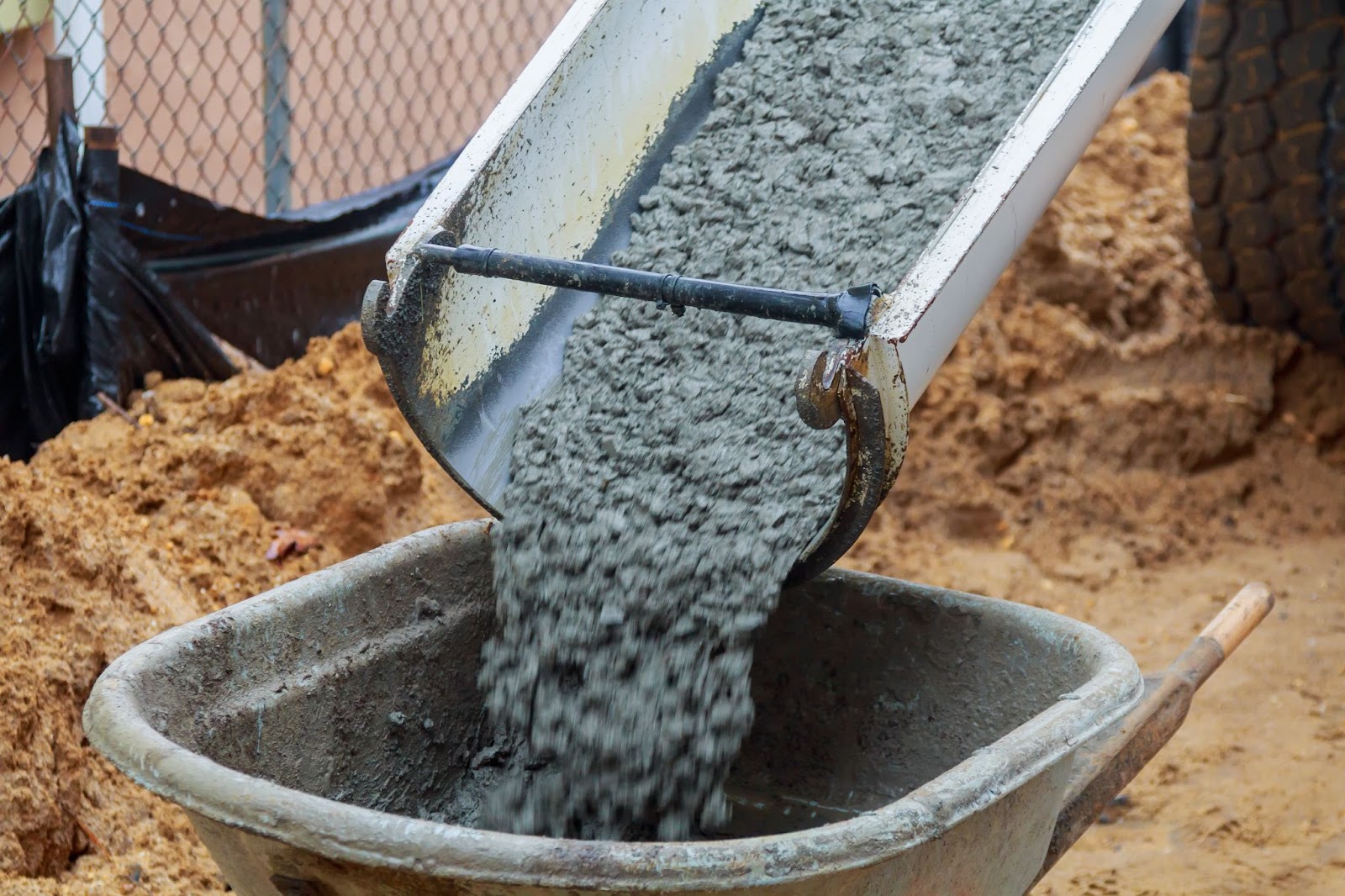 A wheelbarrow filled with cement from a mixer at a construction site, with brown soil visible in the background.
