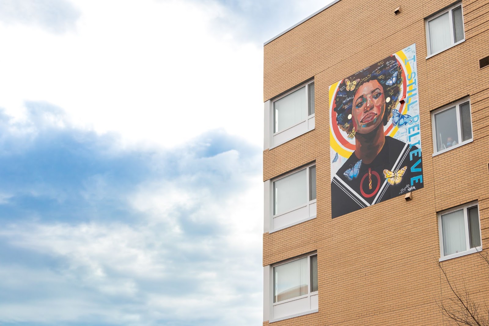  An apartment building surrounded by blue, slightly cloudy skies. On the building is a mural–a portrait of a black woman surrounded by a halo of light and butterflies, with the text I STILL BELIEVE running along the side of the mural.