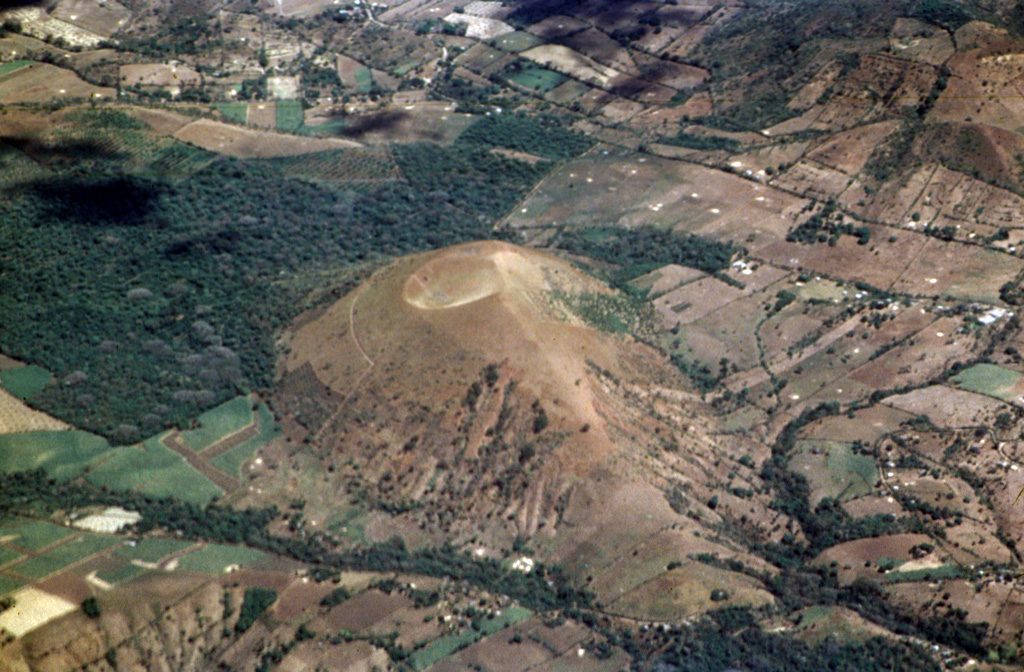 An aerial view of Chingo volcano, an extinct volcano in El Salvador, surrounded by patchwork fields and forested areas under a clear sky.