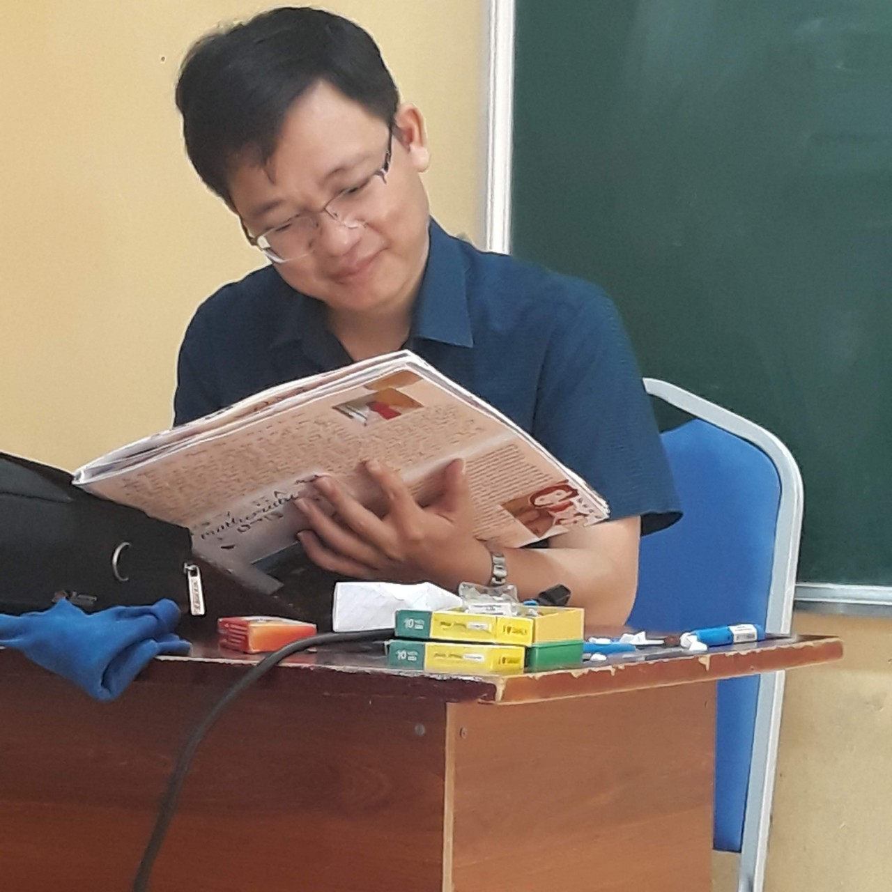 A person reading a newspaper in front of a chalkboardDescription automatically generated