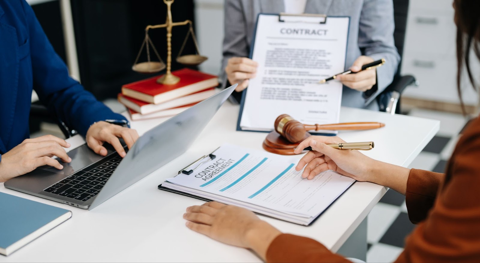 A cropped shot of two business lawyers discussing legal compliance with a female entrepreneur. On the desk are the scales of justice atop three red books, a laptop, a gavel, and various documents.