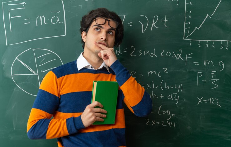 Thoughtful young geometry teacher wearing glasses on forehead standing in front of chalkboard in classroom holding closed book