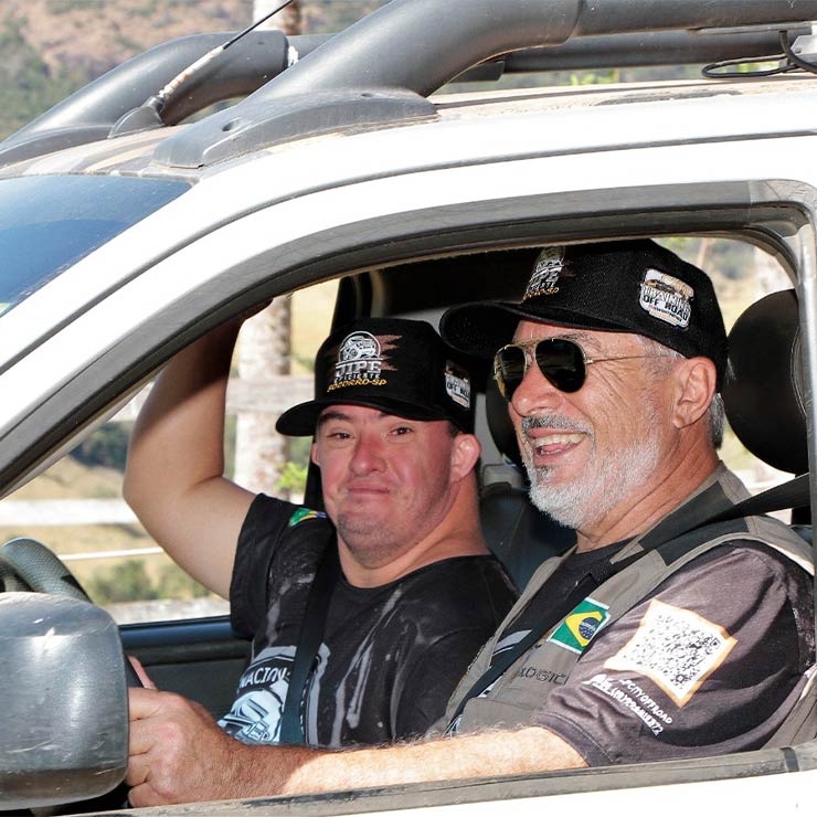 Dois homens sorrindo dentro de um jipe. Ambos usam bonés e camisetas temáticas de um evento off-road.