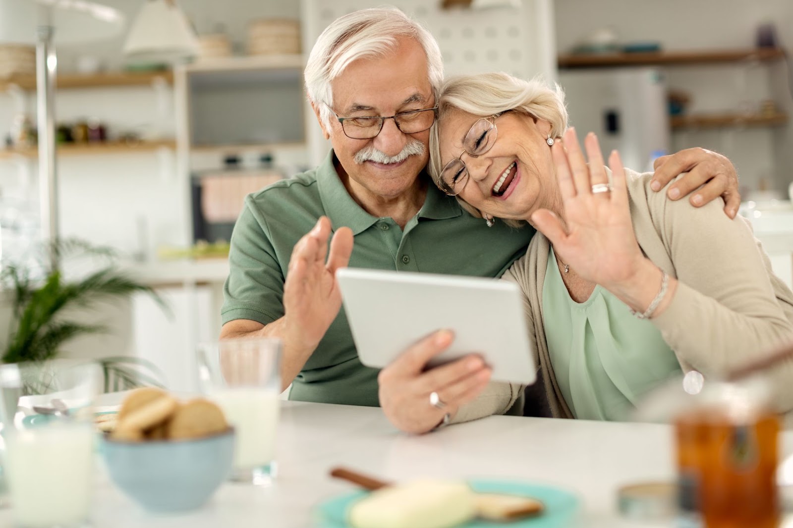 An elderly couple smile and wave at someone during a video call on a tablet.