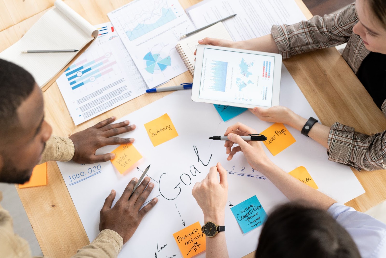 A top shot of three diverse business people collaborating on a business plan. One person holds a tablet, while the table is covered with various illustrations and documents, highlighting their teamwork and strategic planning.