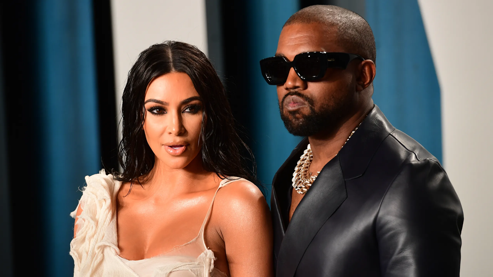 Kim Kardashian y Kanye West posando juntos en un evento, Kim lleva un vestido blanco de un solo hombro y Kanye lleva un traje de cuero negro y lentes de sol.