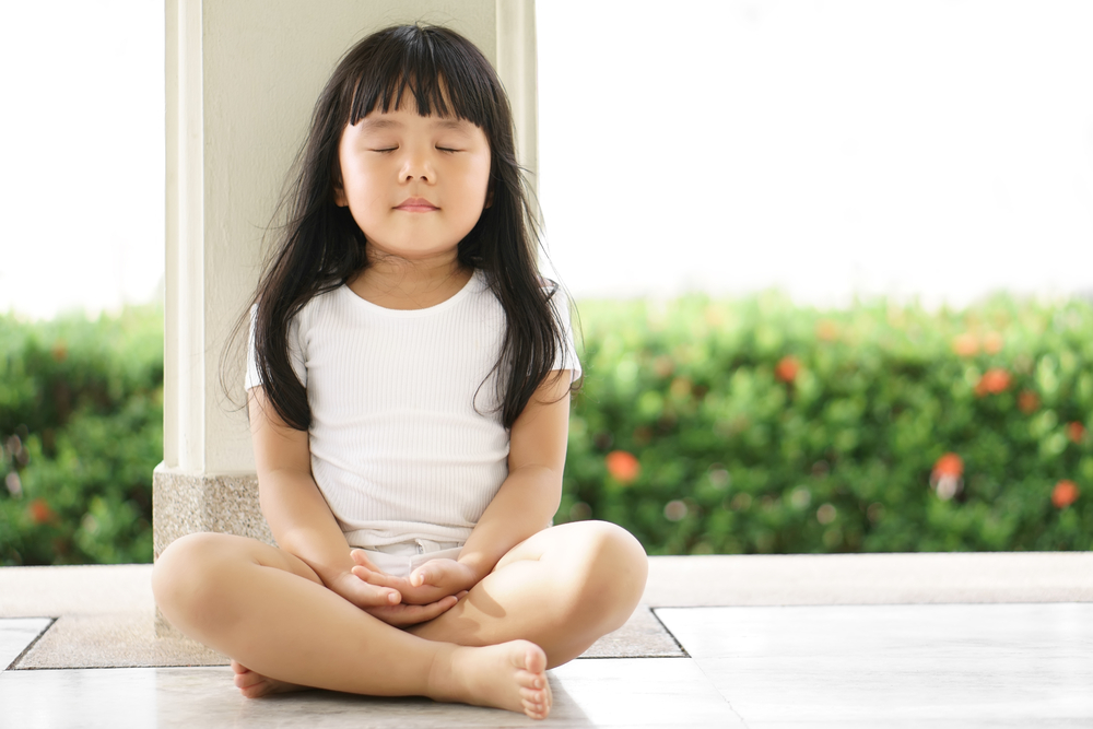 A little kid girl sitting in a meditation pose by the garden, engaging in spiritual wellness activities from a young age.