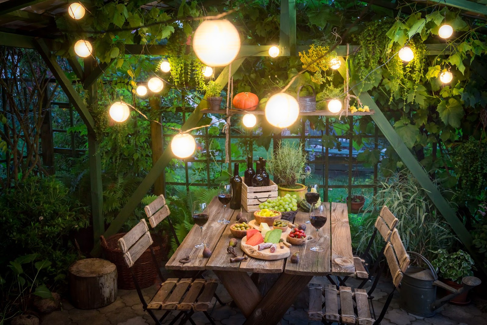 An outdoor wooden dining table is elegantly set for an evening dinner, adorned with a spread of cheeses, fruits, and a glass of red wine, creating a charming atmosphere for outdoor dining.