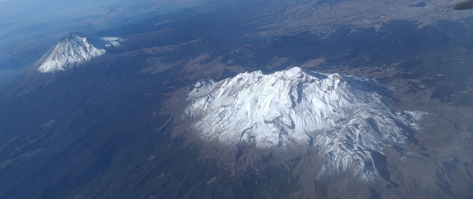 Aerial view of two snow-capped mountains with rugged terrain, showcasing their majestic peaks against a backdrop of forested slopes.