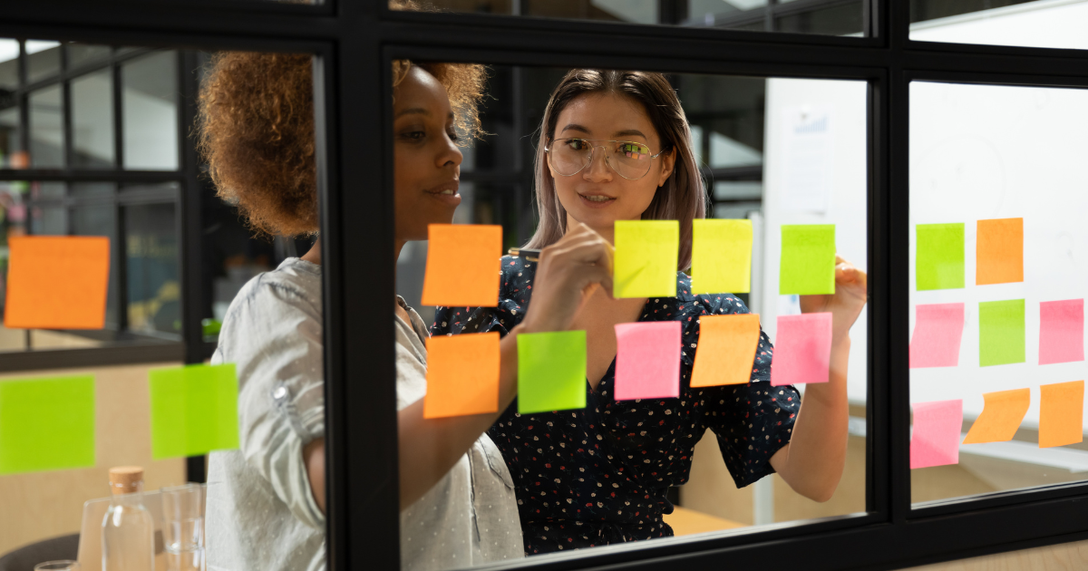 A diverse group of two professionals standing together in a modern office, writing on sticky notes, and smiling confidently, showcasing the benefits of Scrum Master training.
