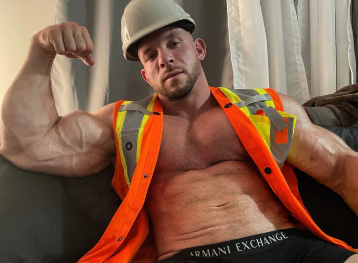 Cody Mac wearing a construction hard hat shirtless wearing an orange safety vest while flexing his massive biceps in Armani Exchange underwear
