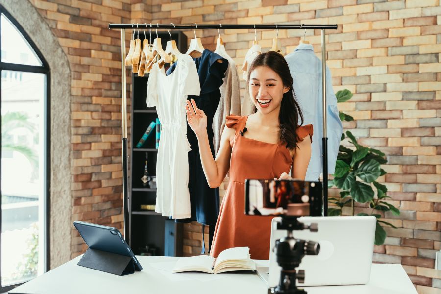 Woman live streaming fashion content with clothing rack in the background.