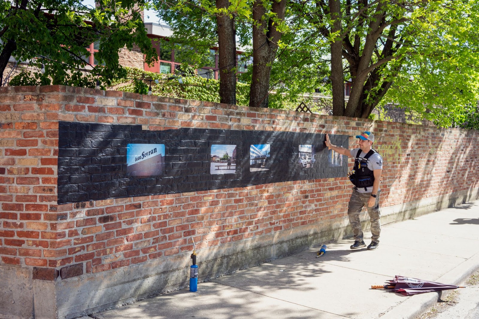 Image: Installation view, RER B-ANLIEUS,2021. A man just finished adhering a poster of Assia Labbas’ RER B-ANLIEUS onto a brick wall along the outer perimeter of Experimental Station, Woodlawn. The piece looks like a strip of film for a Kodak carousel projector, with vignette-style photographs from different locations in Paris along the RER-B train line as the slides. Courtesy of Villa Albertine.