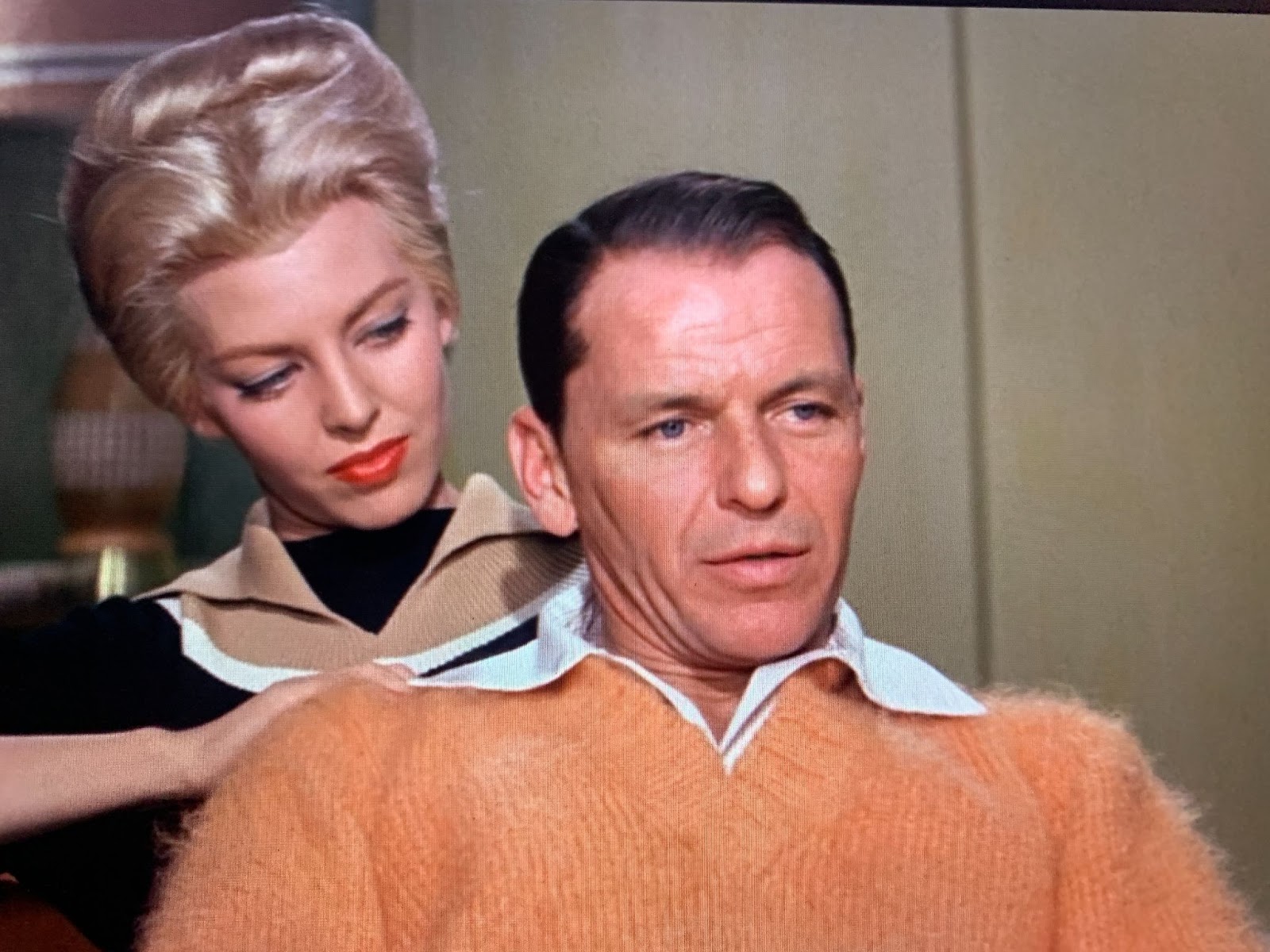 Frank Sinatra, shown from the chest up, sits in the center of the frame facing slightly to his left, or the viewer's right. He wears a shirt with a wide, floppy white collar under an orange sweater with a thickly cabled V-neck and a knit so fuzzy that a nimbus of orange fluff outlines him. Behind him and rubbing his right shoulder is a woman in a bleached-blonde beehive with blue eye shadow and bright red lipstick, wearing a dark sweater with a tan collar and yoke outlined in white. 