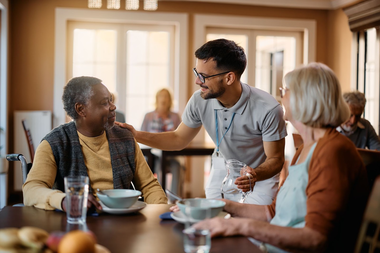 A dining room staff member at an assisted living community serves water to a group of residents enjoying a meal.