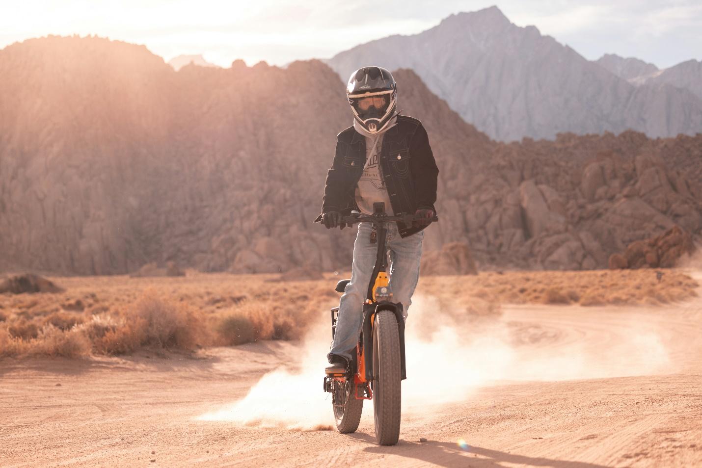 a person on an ebike riding on a rough terrain . The rider is wearing an helmet, black jacket and jeans. There is sand on the ground and mountains in the background.