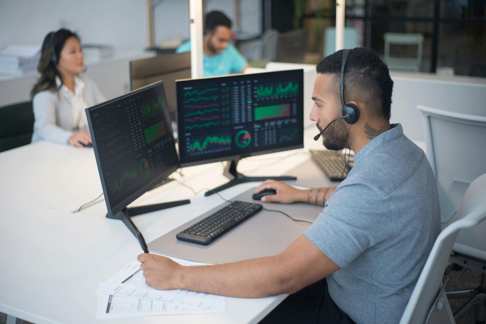 A man monitors data on dual computer screens with colleagues in the background.