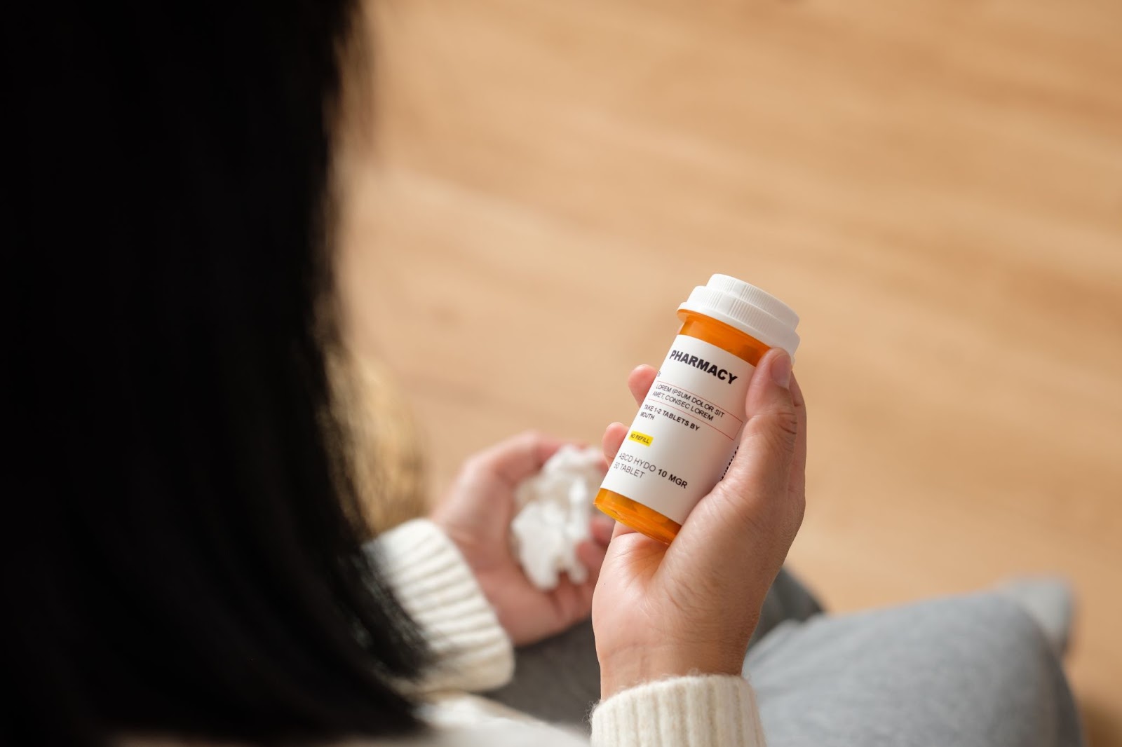 A cropped view of a woman with dark hair holding a prescription bottle of pills in one hand and holding a tissue in the other hand.