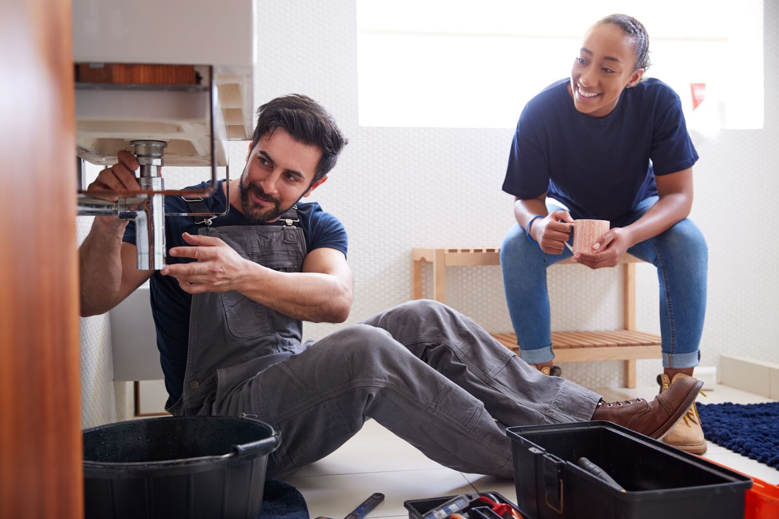 A bearded male plumber in black shirt and denim overalls teaches a seated female apprentice how to fix a leaking sink in a home bathroom.