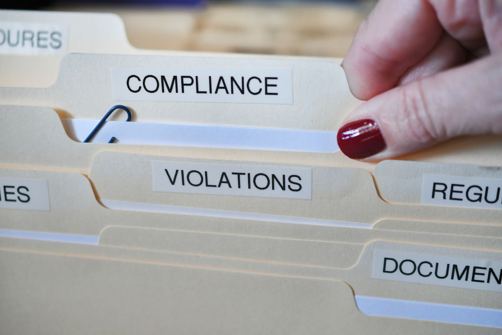 A lady’s hand, covered in red nail polish, removes a 'Compliance' folder from an office filing cabinet amidst other files and folders, possibly for a GRC meeting.