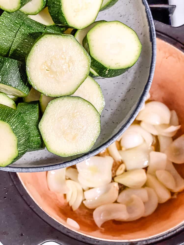 a bowl of sliced zucchini being held above a pot with onions and garlic.