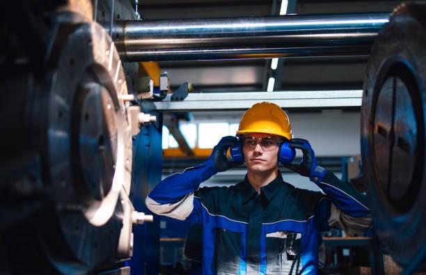 A young male worker in a factory wearing a safety helmet and ear protection stands next to heavy machinery, emphasizing industry safety and professionalism. Focused Factory Worker Adjusts Safety Gear in Industrial Setting precision manufacturing stock pictures, royalty-free photos & images