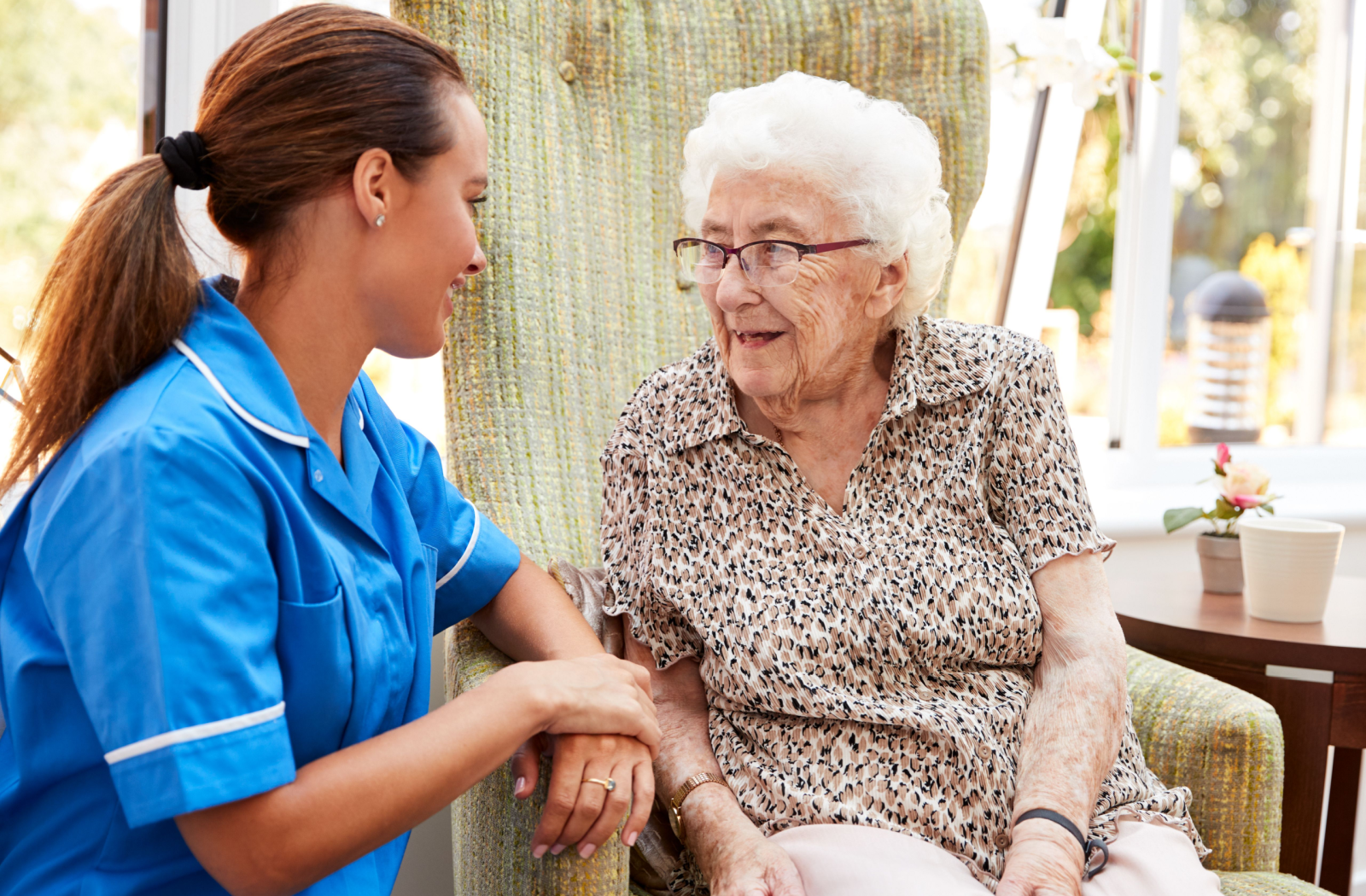 A nurse leans on the chair arm of a senior person as they smile at each other in a well-lit dining room