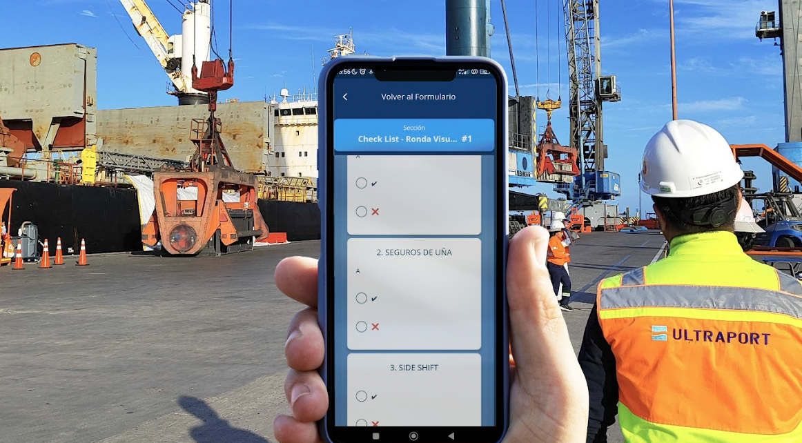  Person holding a cell phone with a port and workers in vests and hard hats in the background. 