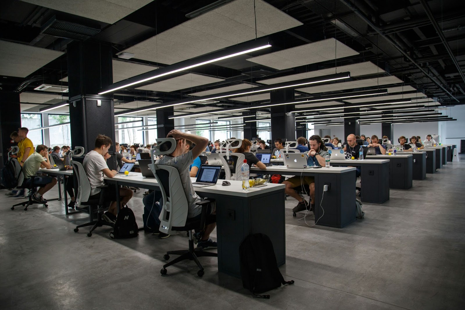 A large open-plan office with many people working at long rows of desks, each using a laptop.