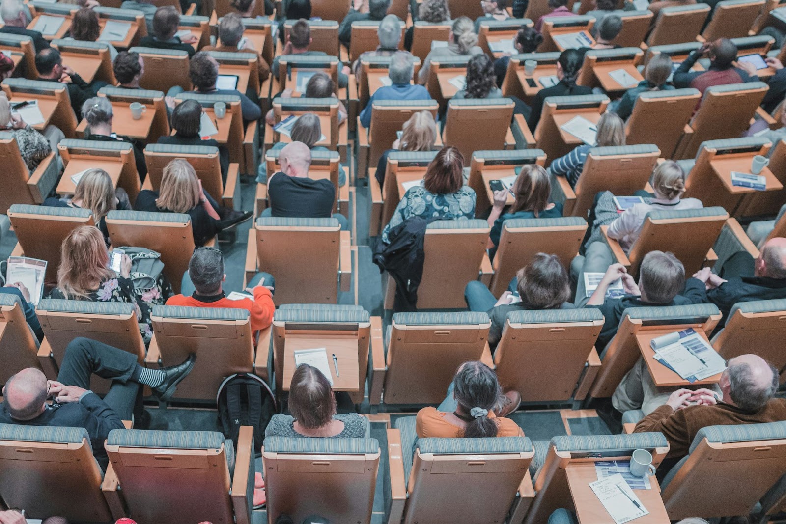 A large group of people seated in rows of lecture hall chairs, viewed from above, attentively listening to a presentation.