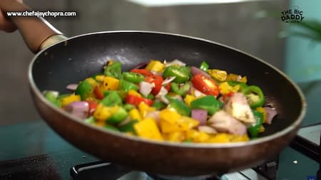 A pan with diced vegetables like yellow and green capsicums, onions, and red chilies being sautéed.