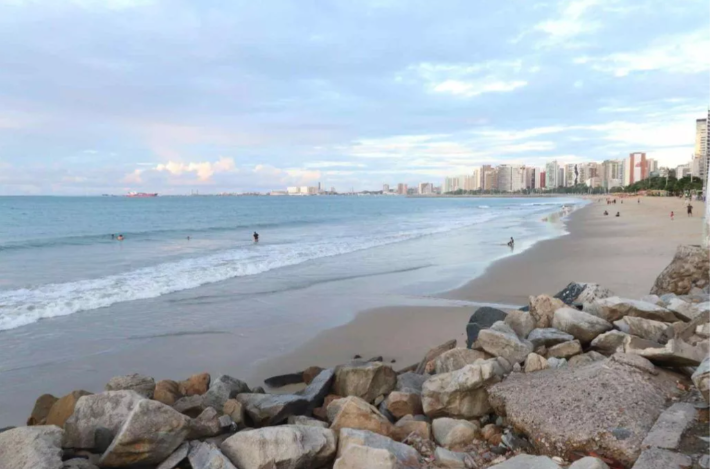 Praia de Iracema, Fortaleza. A areia é de uma coloração marrom clara e se encontra com um mar azul acinzentado praticamente sem ondas. Ao fundo, aparecem os prédios da cidade.