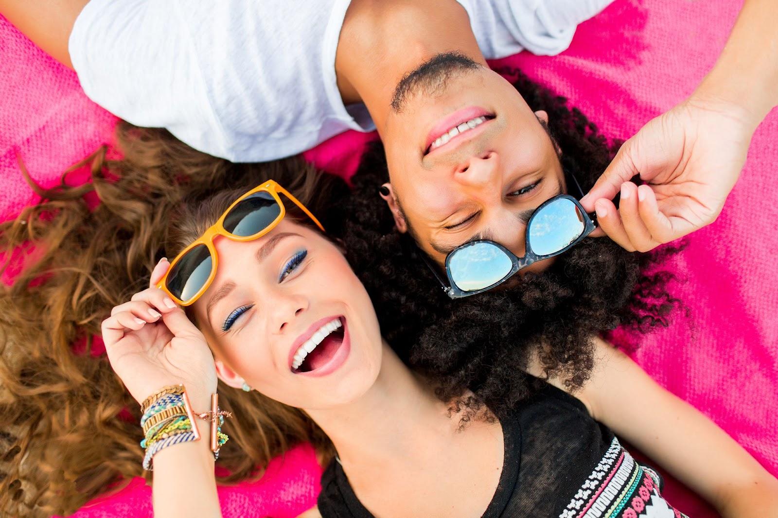 View from above of 2 young people laying on a pink blanket and holding their sunglasses above their eyes and smiling.