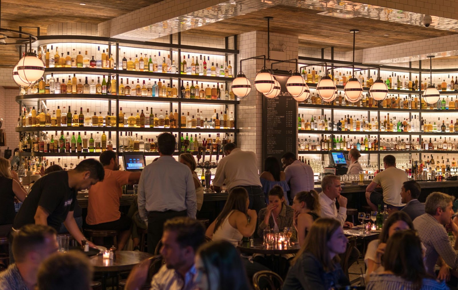 A busy bar in the evening, customers huddled over tables and queuing for drinks. The bar shelves are heavily stocked with bottles.