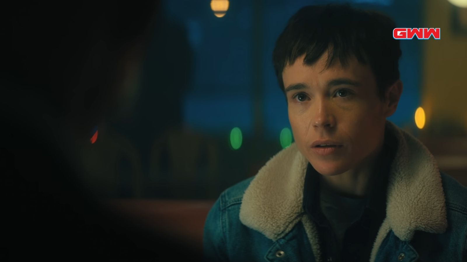 Vanya with short hair in a denim jacket, talking in a dimly lit diner.