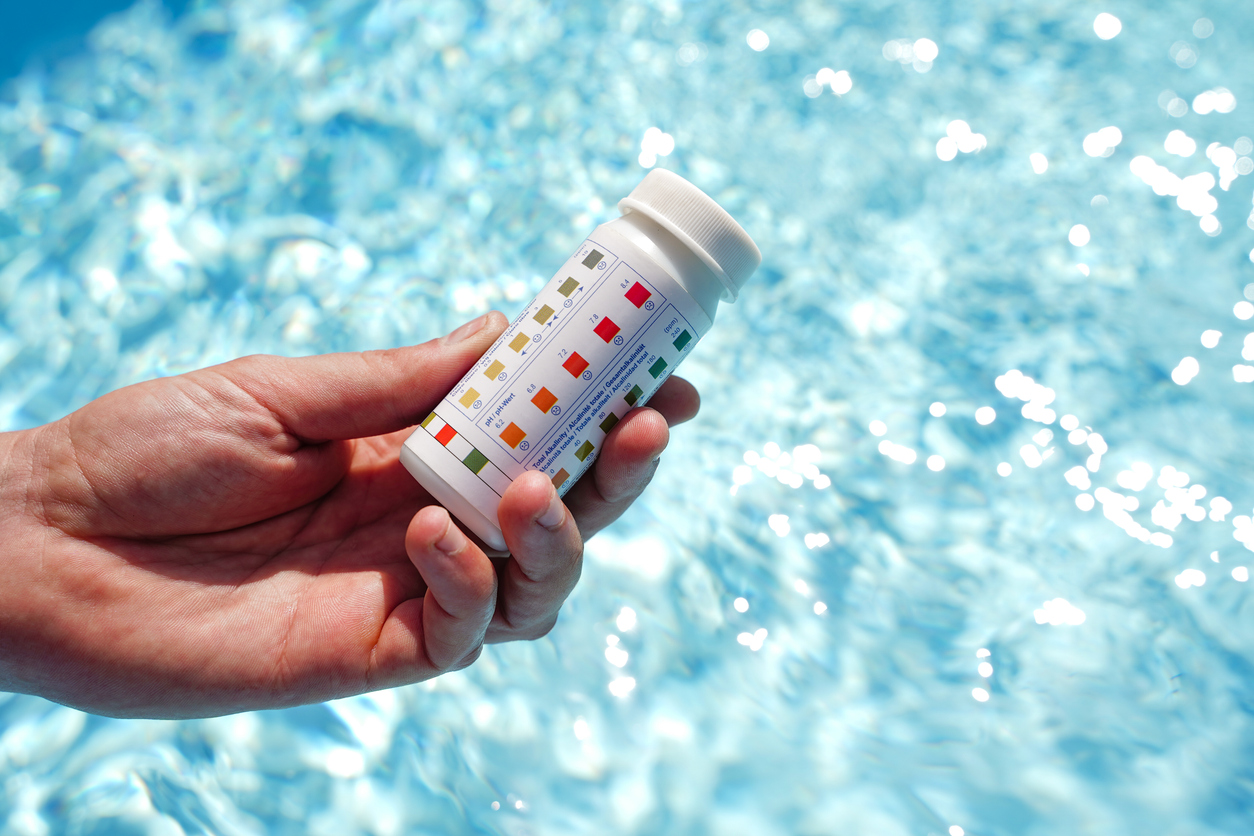 A close-up of a swimming pool water pH and chemical balance test, which is held above a pool. 