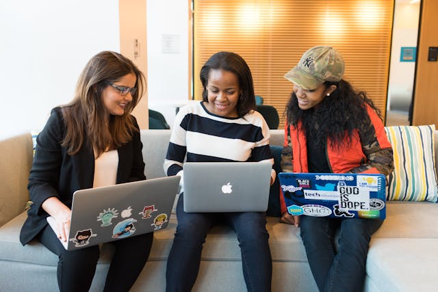 Three women sitting on a couch with laptops, working together in a cozy living room setting.