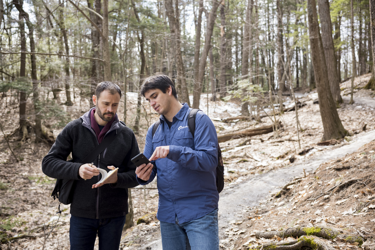 Two men in the woods holding a notebook and a phone. 