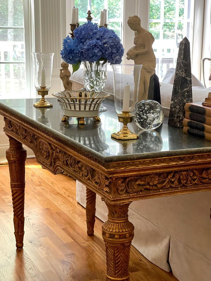 Ornately carved gilt table with dark marble top, styled with crystal vase, blue hydrangeas, brass candlesticks, classical statue, glass orb, and antique books. Natural light highlights the details.