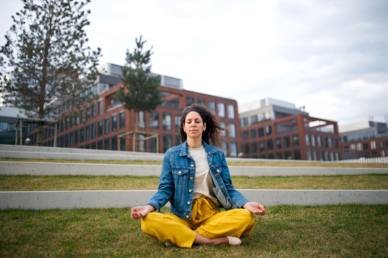 Woman outside meditating on a lawn with buildings in the background. 