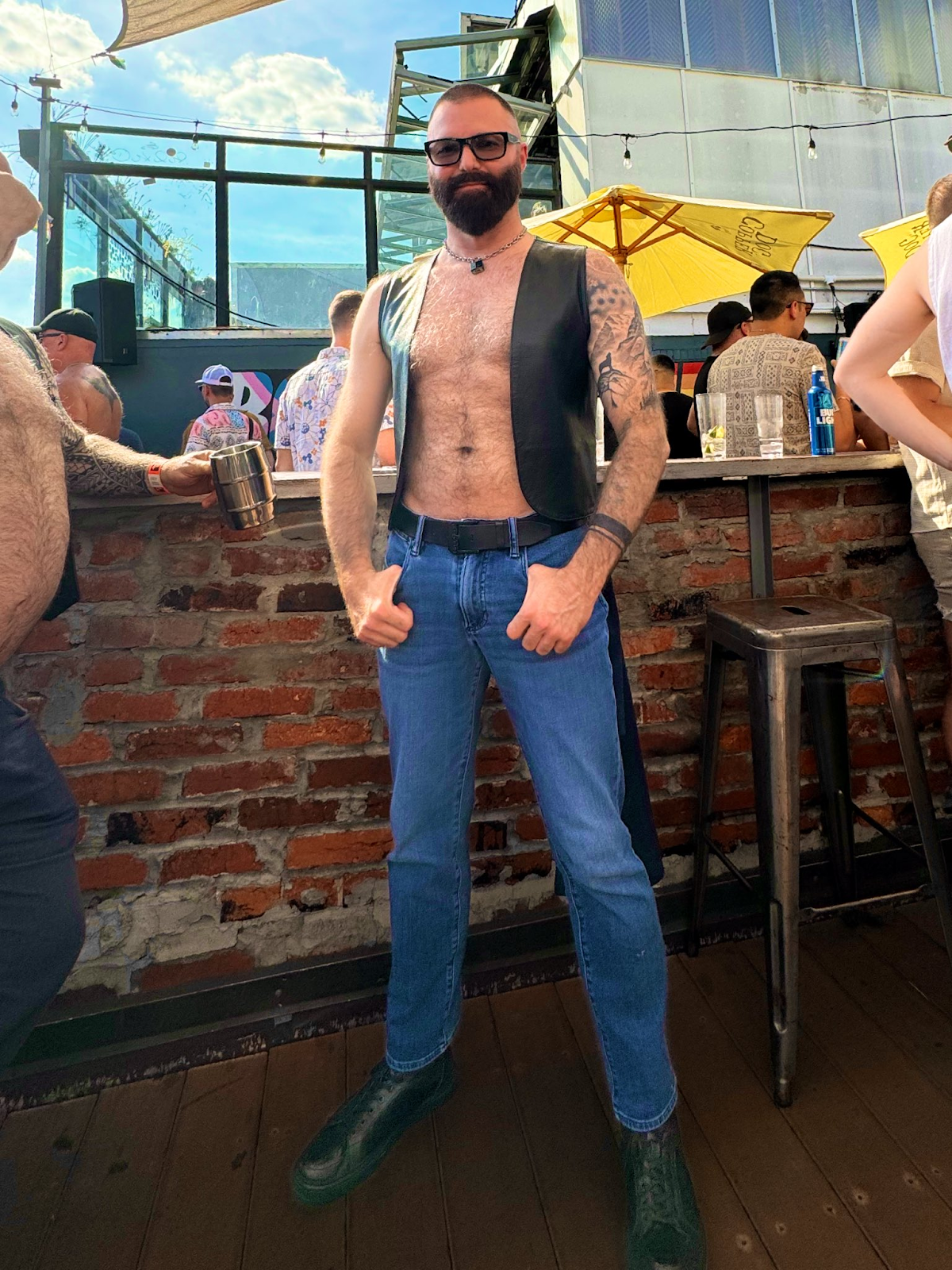 Rob Montana muscle bear posing outside at gay leather event in leather kink vest with hairy muscle bears drinking and partying around him outdoors