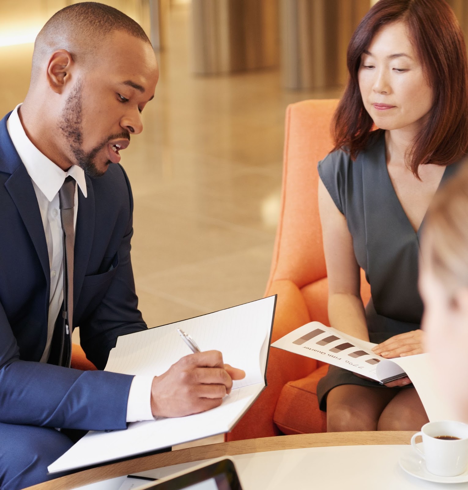 A male business consultant in a business suit discusses and writes his recommendations to his female Asian client seated on an orange-cushioned chair. A third person is half shown, and a cup of coffee is on the table.