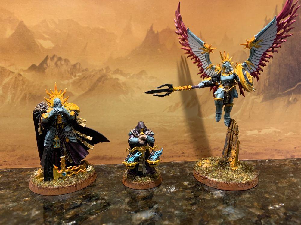 Preview miniatures of an armored warrior with an axe, his squire bearing a censer, and a winged warrior with a trident. Painted in blue and gold metal with purple cloth.