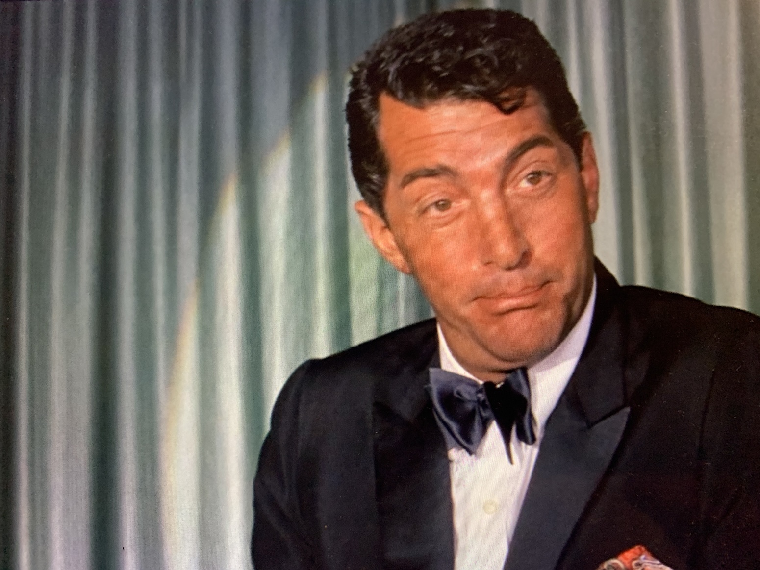 Dean Martin, tanned, shown from mid-chest up in a tuxedo with black bow tie, cocking an eyebrow and wrinkling his mouth. The edge of the spotlight shining on him shows on the gray-green curtain hanging behind him. 