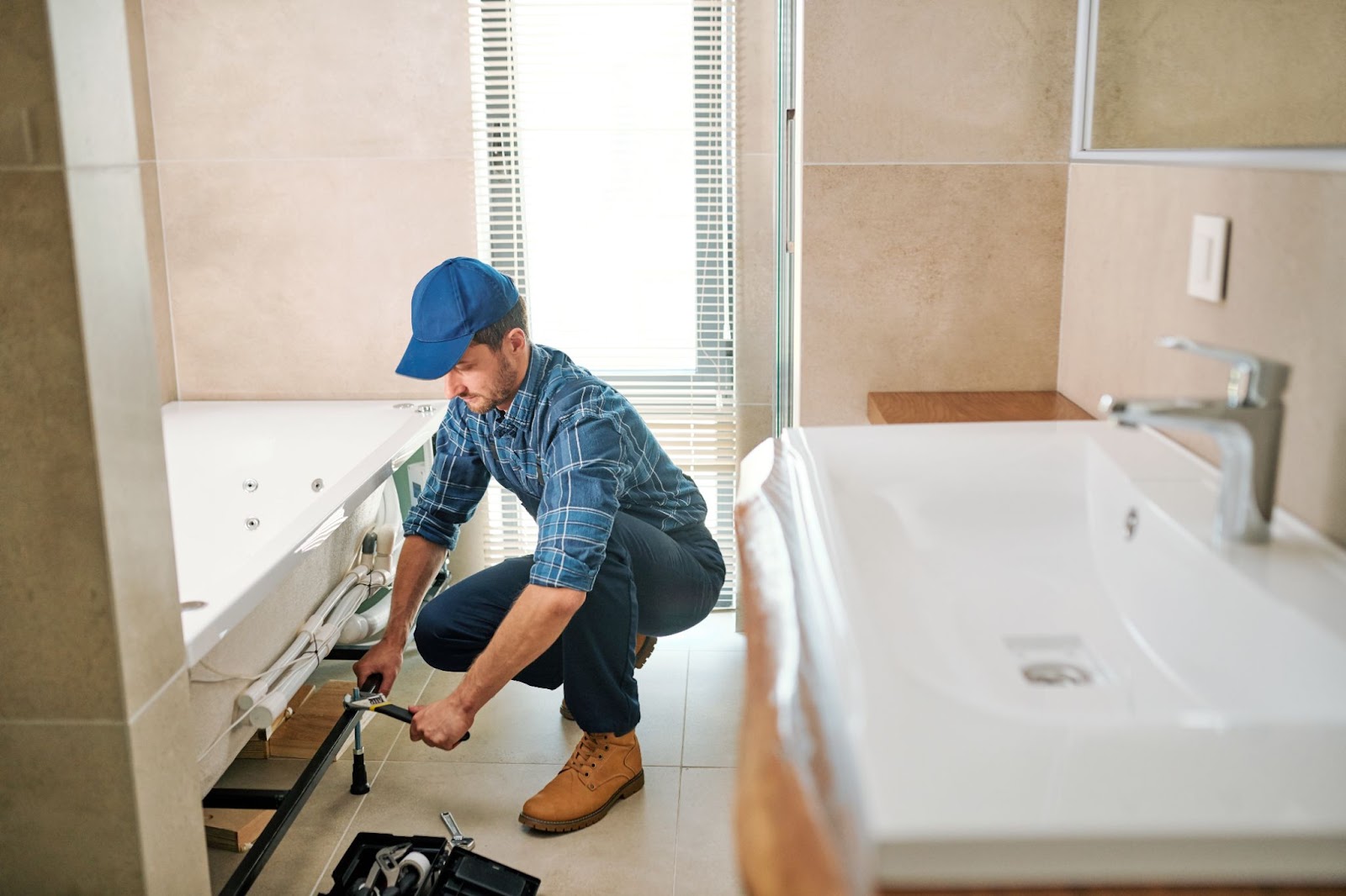 A young worker in uniform crouches while fixing a detail on a bathtub. A white ceramic sink is visible to the right, and a window with blinds is in the background.