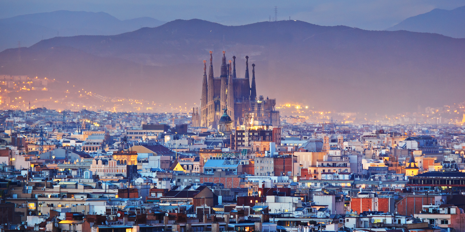 A stunning panoramic view of Barcelona at dusk, featuring the iconic Sagrada Familia church prominently in the center, surrounded by the city's urban landscape. The distant mountains add a picturesque backdrop, while the city's lights begin to illuminate the skyline, creating a beautiful and serene evening atmosphere.